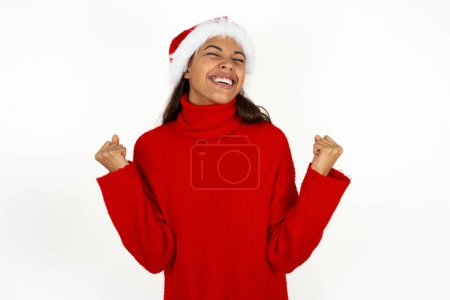 Téléchargez les photos : Jeune belle femme portant un pull rouge et santa claus chapeau à Noël célébrant surpris et étonné pour le succès avec les bras levés et les yeux fermés. Concept gagnant. - en image libre de droit