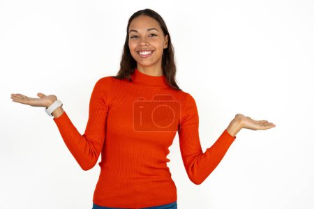 Foto de Alegre alegre optimista joven hispana vistiendo cuello alto rojo sobre fondo blanco sosteniendo dos palmeras espacio de copia - Imagen libre de derechos