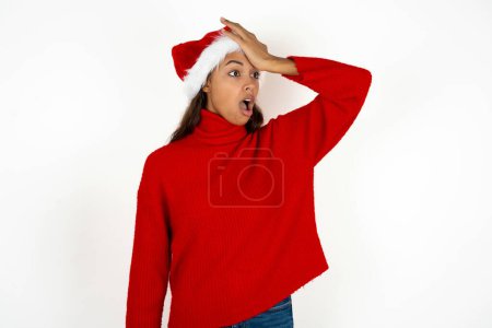 Foto de Joven hermosa mujer vistiendo suéter rojo y sombrero de Santa Claus en Navidad sorprendida con la mano en la cabeza por error, recordar el error. Olvidado, mal concepto de memoria. - Imagen libre de derechos