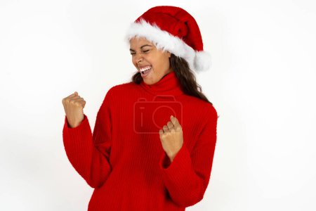 Foto de Joven hermosa mujer vistiendo suéter rojo y sombrero de Santa Claus en Navidad muy feliz y emocionado haciendo gesto ganador con los brazos levantados, sonriendo y gritando por el éxito. Concepto de celebración. - Imagen libre de derechos