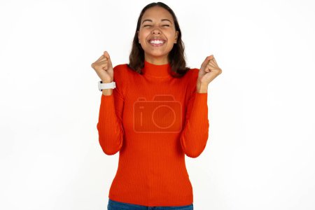 Téléchargez les photos : Jeune femme hispanique portant col roulé rouge sur fond blanc étant excité pour le succès avec les bras levés et les yeux fermés célébrant la victoire. Concept gagnant. - en image libre de droit