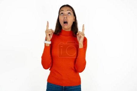 Téléchargez les photos : Jeune femme hispanique portant col roulé rouge sur fond blanc étonné et surpris regardant vers le haut et pointant avec les doigts et les bras levés. - en image libre de droit