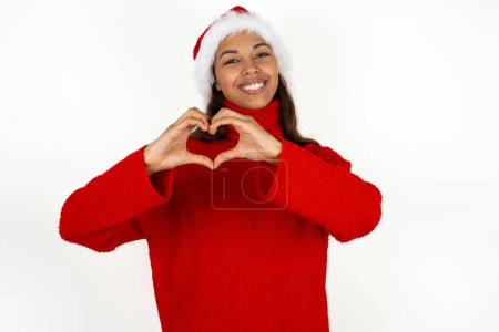 Téléchargez les photos : Jeune belle femme portant un pull rouge et santa claus chapeau à Noël souriant dans l'amour faisant forme de symbole de coeur avec les mains. Concept romantique. - en image libre de droit