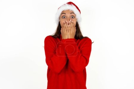 Foto de Vivaz joven hermosa mujer vistiendo suéter rojo y sombrero de Santa Claus en la Navidad risitas alegremente, cubre la boca, tiene risa natural, escucha historia positiva o anécdota divertida - Imagen libre de derechos
