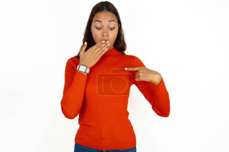 Téléchargez les photos : Choqué jeune femme hispanique portant col roulé rouge sur fond blanc regarder étonnamment vers le bas, indique à blanc avec le doigt avant, modèle effrayé - en image libre de droit