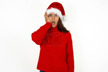 Téléchargez les photos : Embarrassé jeune belle femme portant un pull rouge et santa claus chapeau à Noël avec une expression choquée, exprime une grande stupéfaction, pose modèle perplexe intérieur - en image libre de droit