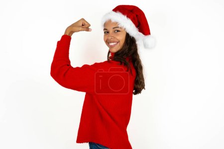 Photo for Portrait of powerful cheerful young beautiful woman wearing red sweater and santa claus hat at christmas showing muscles. - Royalty Free Image