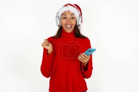 Téléchargez les photos : Une belle jeune femme positive portant un pull rouge et un chapeau santa claus à Noël tient un téléphone portable moderne connecté à un casque, serre le poing de bonnes émotions, s'exclame avec joie, - en image libre de droit