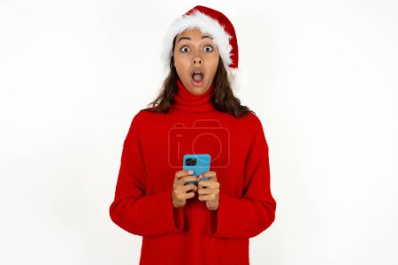 Photo for Shocked young beautiful woman wearing red sweater and santa claus hat at christmas opens mouth hold phone reading advert unbelievable big shopping prices - Royalty Free Image