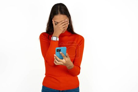 Photo for Young hispanic woman wearing red turtleneck over white background looking at smartphone feeling sad holding hand on face. - Royalty Free Image