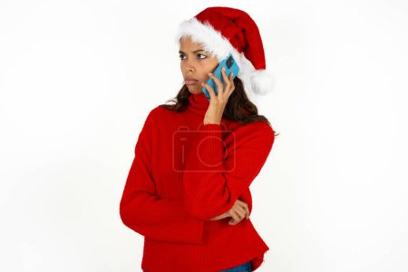 Téléchargez les photos : Triste jeune belle femme portant un pull rouge et un chapeau santa claus à Noël parlant sur smartphone. Concept de communication. - en image libre de droit
