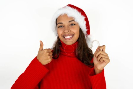 Photo for Young beautiful woman wearing red sweater and santa claus hat at christmas holding an invisible braces aligner and rising thumb up, recommending this new treatment. Dental healthcare concept. - Royalty Free Image
