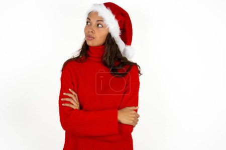 Téléchargez les photos : Agréable jeune belle femme portant un pull rouge et santa claus chapeau à Noël garde les mains croisées sur la poitrine semble heureusement de côté - en image libre de droit