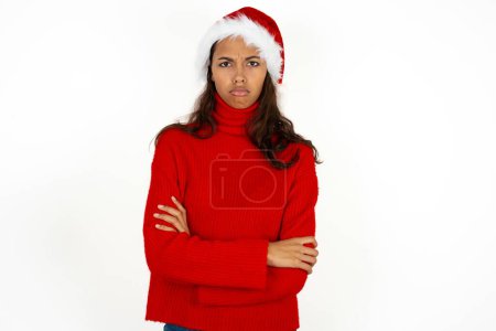 Foto de Sombría insatisfecha joven hermosa mujer vistiendo suéter rojo y sombrero de Santa Claus en la Navidad se ve con expresión miserable en la cámara de debajo de la frente, hace infeliz mueca - Imagen libre de derechos