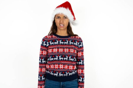 Photo for Portrait of dissatisfied beautiful hispanic woman wearing knitted sweater and santa claus hat over white background smirks face, purses lips and looks with annoyance at camera, discontent hearing something unpleasant - Royalty Free Image