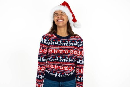 Photo for Positive beautiful hispanic woman wearing knitted sweater and santa claus hat over white background with overjoyed expression closes eyes and laughs shows white perfect teeth - Royalty Free Image