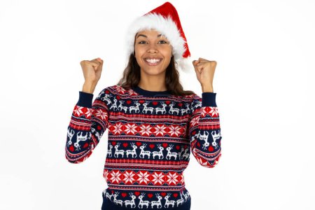 Photo for Shocked ecstatic beautiful hispanic woman wearing knitted sweater and santa claus hat over white background win luck lottery raise hands up shout yea - Royalty Free Image