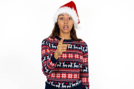 Photo for Shocked beautiful hispanic woman wearing knitted sweater and santa claus hat over white background points at you with stunned expression - Royalty Free Image