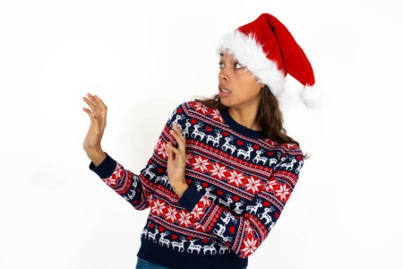 Photo for Displeased beautiful hispanic woman wearing knitted sweater and santa claus hat over white background keeps hands towards empty space and asks not come closer sees something unpleasant - Royalty Free Image