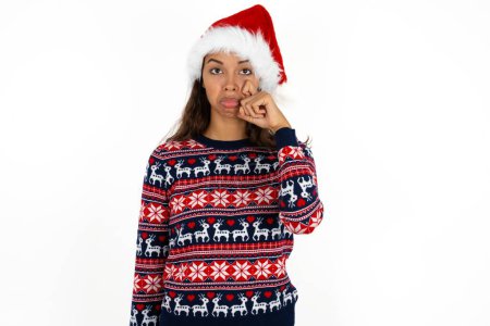 Photo for Disappointed dejected beautiful hispanic woman wearing knitted sweater and santa claus hat over white background wipes tears stands stressed with gloomy expression. Negative emotion - Royalty Free Image