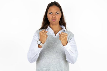 Foto de Displeased annoyed young beautiful hispanic woman over white background clenches fists, gestures pissed, ready to revenge, looks with aggression at camera stands full of hate, being pressured - Imagen libre de derechos