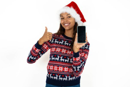 Foto de Hermosa mujer hispana con suéter de punto y sombrero de Santa Claus sobre fondo blanco Mostrar pantalla en blanco smartphone, pulgar hacia arriba recomendar nueva aplicación - Imagen libre de derechos