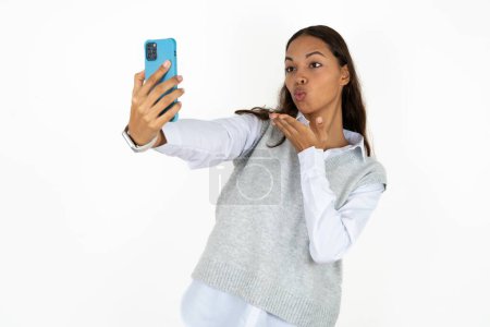 Photo for Young beautiful hispanic woman over white background blows air kiss at camera of smartphone and takes selfie, sends mwah via online call. - Royalty Free Image