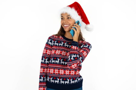 Photo for Pleasant looking happy beautiful hispanic woman wearing knitted sweater and santa claus hat over white background has nice telephone conversation and looks aside, has nice mood and smiles positively while talks via cell phone - Royalty Free Image