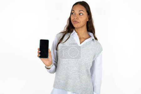 Téléchargez les photos : Jeune belle femme hispanique sur fond blanc tient un nouveau téléphone mobile et semble mystérieux de côté montre affichage vierge de cellulaire moderne - en image libre de droit