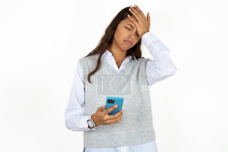 Photo for Upset depressed young beautiful hispanic woman over white background makes facepalm as forgot about something important holds mobile phone expresses sorrow and regret blames - Royalty Free Image