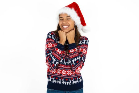 Foto de Alegre hermosa mujer hispana con suéter de punto y sombrero de Santa Claus sobre fondo blanco tiene expresión satisfecha tímida, sonríe ampliamente, muestra dientes blancos, emociones de la gente - Imagen libre de derechos