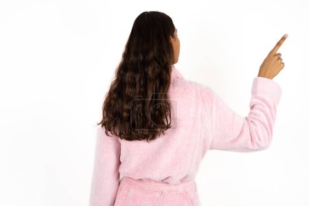 Téléchargez les photos : Jeune femme portant un peignoir rose sur fond blanc pointant vers l'objet sur l'espace de copie, vue arrière. Tourne le dos - en image libre de droit