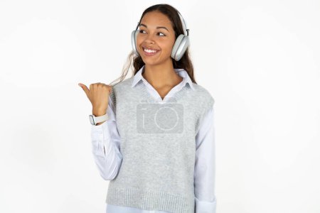 Photo for Young beautiful hispanic woman over white background listens audio track via wireless headphones points thumb away advertises copy space - Royalty Free Image