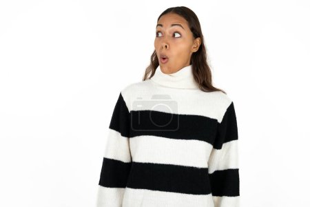 Foto de Joven mujer hermosa impactada vistiendo suéter a rayas sobre fondo blanco mira espacio vacío con la boca abierta gritando: ¡Oh Dios mío! No puedo creerlo.. - Imagen libre de derechos