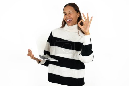 Téléchargez les photos : Attrayant joyeux qualifié jeune belle femme portant un pull rayé sur fond blanc en utilisant un ordinateur portable montrant ok-signe winkin - en image libre de droit