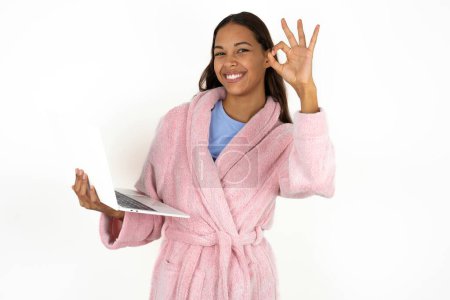 Foto de Mujer joven positiva con albornoz rosa sobre fondo blanco mantenga los dedos inalámbricos netbook mano muestran símbolo okey - Imagen libre de derechos