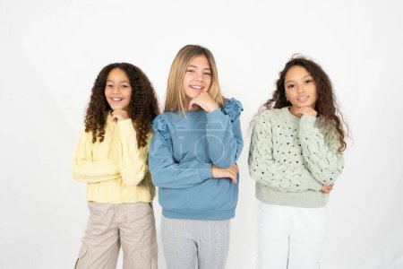 Optimistic Three young beautiful multiracial kid girls keeps hands partly crossed and hand under chin, looks at camera with pleasure.