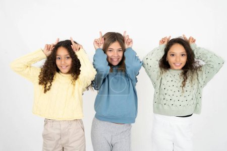 Photo for Funny Three young beautiful multiracial kid girls shows horns, fingers on head gesture, posing silly and cute - Royalty Free Image