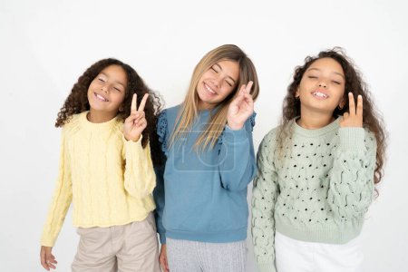 Foto de Tres jóvenes hermosas niñas multirraciales sonriendo con la cara feliz guiñando el ojo a la cámara haciendo signo de victoria. Número dos.. - Imagen libre de derechos