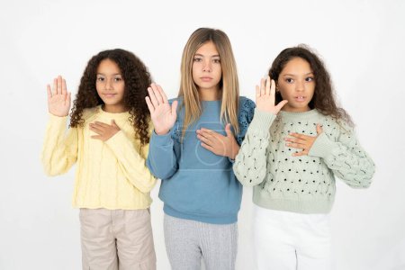 Drei junge schöne multirassische Mädchen, die mit der Hand auf der Brust und der offenen Handfläche schwören und einen Treueschwur leisten