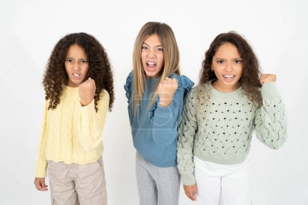 Three young beautiful multiracial kid girls angry and mad raising fist frustrated and furious while shouting with anger. Rage and aggressive concept.