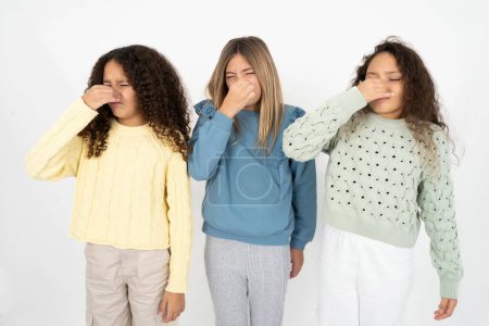 Photo for Three young beautiful multiracial kid girls smelling something stinky and disgusting, intolerable smell, holding breath with fingers on nose. Bad smell - Royalty Free Image