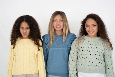 Photo for Three young beautiful multiracial kid girls keeps teeth clenched, frowns face in dissatisfaction, irritated because of much duties. - Royalty Free Image