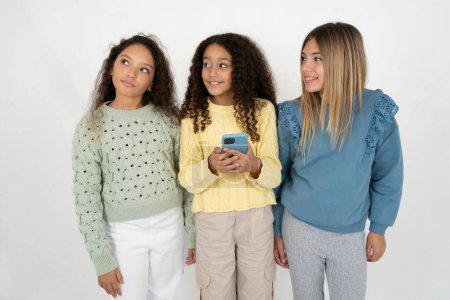 Téléchargez les photos : Trois jeunes belles jeunes filles multiraciales tenant dans les mains le téléphone mobile et regardant de côté avec un regard rêveur. - en image libre de droit