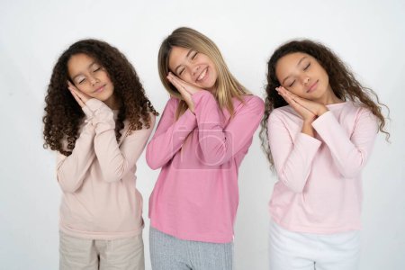Foto de Tres jóvenes hermosas niñas multirraciales se apoya en las palmas prensadas cierra los ojos y tiene una sonrisa agradable sueña con algo - Imagen libre de derechos