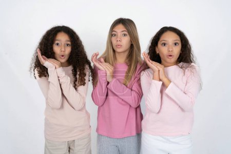 Téléchargez les photos : Surpris émotionnel Trois jeunes belles filles multiraciales enfants frotte les paumes et regarde la caméra avec incrédulité - en image libre de droit
