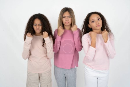 Displeased annoyed Three young beautiful multiracial kid girls clenches fists, gestures pissed, ready to revenge, looks with aggression at camera stands full of hate, being pressured