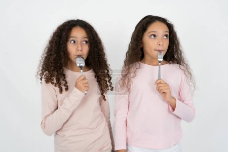 Photo for Photo of dreamy two young beautiful multiracial kid girls lick fork look empty space - Royalty Free Image