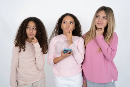 Téléchargez les photos : Trois jeunes belles filles multiraciales enfant pense profondément à quelque chose, utilise le téléphone mobile moderne, essaie de faire un bon message, garde l'index près des lèvres. - en image libre de droit