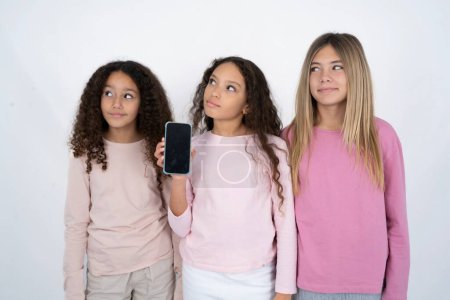 Photo for Three young beautiful multiracial kid girls holds new mobile phone and looks mysterious aside shows blank display of modern cellular - Royalty Free Image